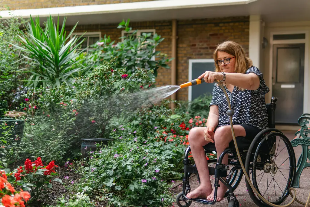 A woman in a wheelchair waters the plants in her front garden.