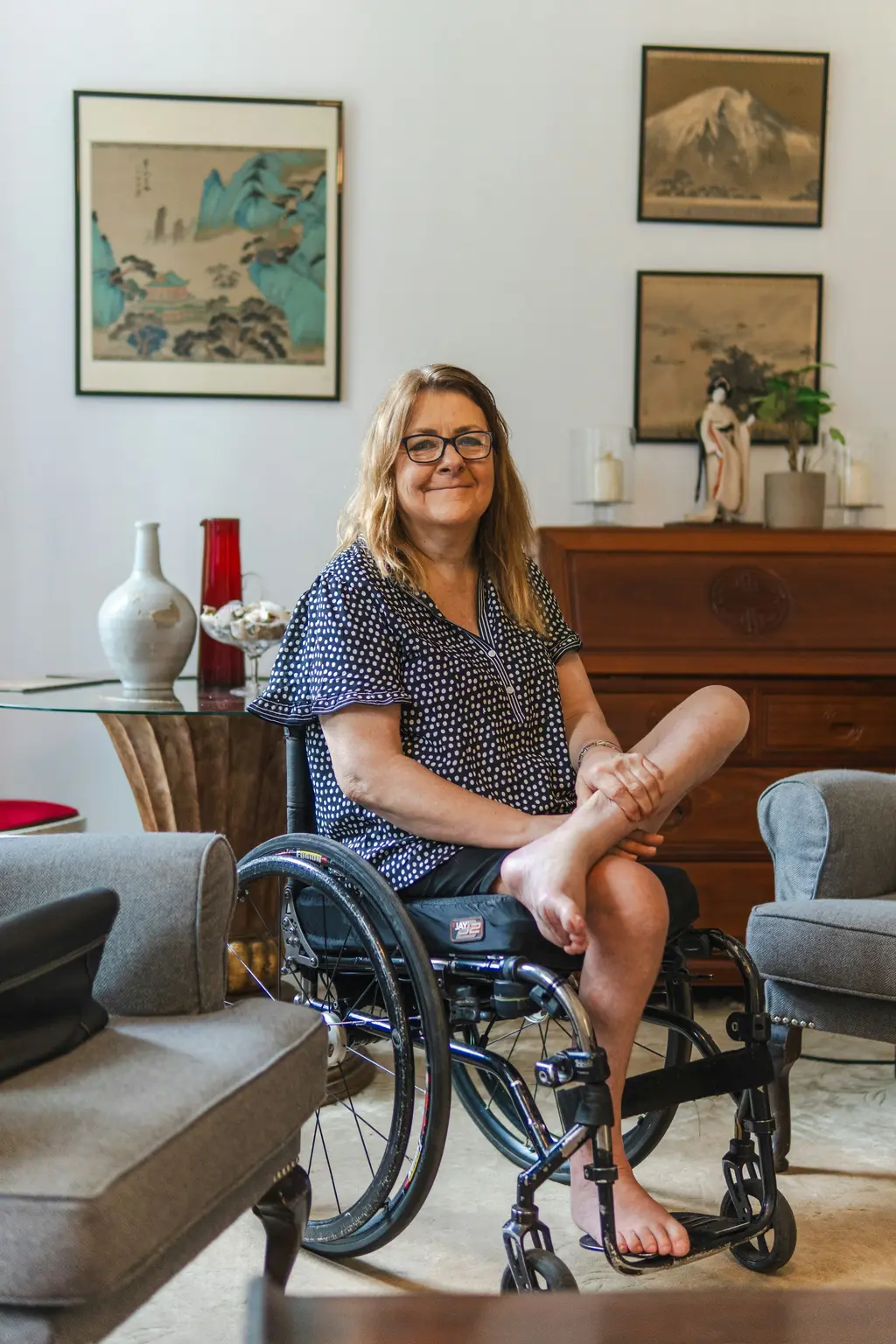 A woman sits in her wheelchair in her living room, smiling, with pictures on the wall behind her.